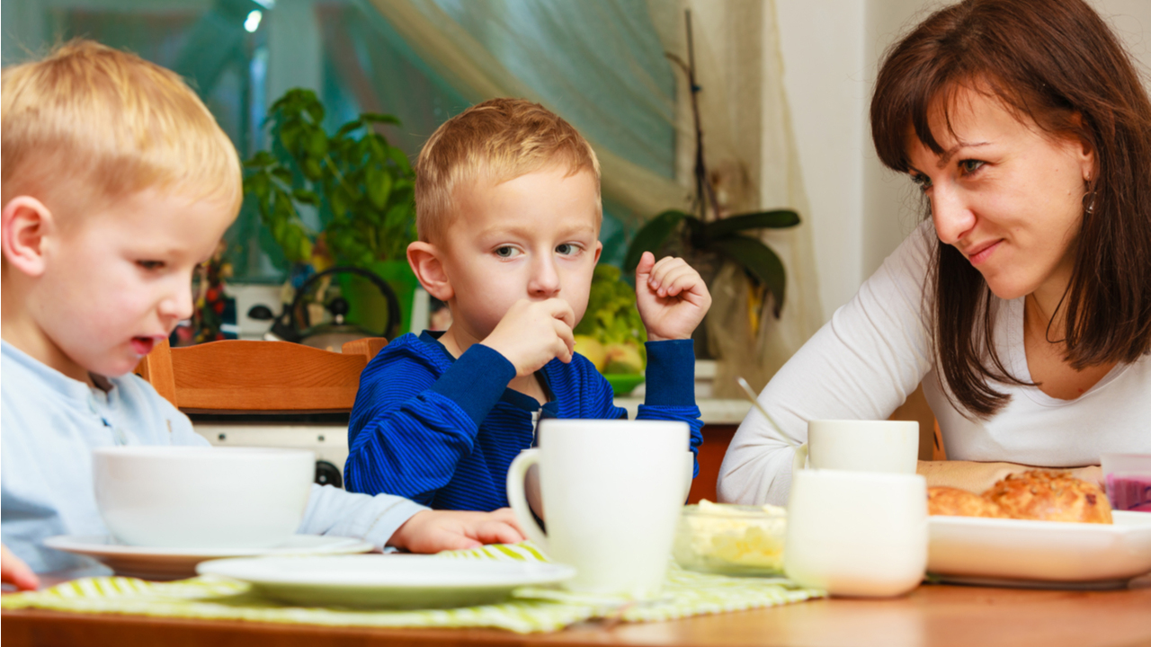 Two young children eat breakfast with mother at the table